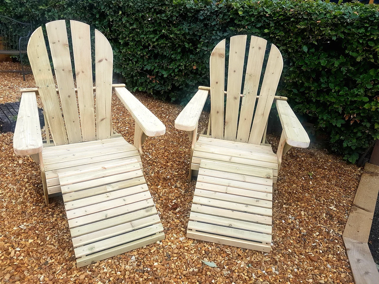 A matching pair of timber Adirondack chairs with slatted footstools on gravel